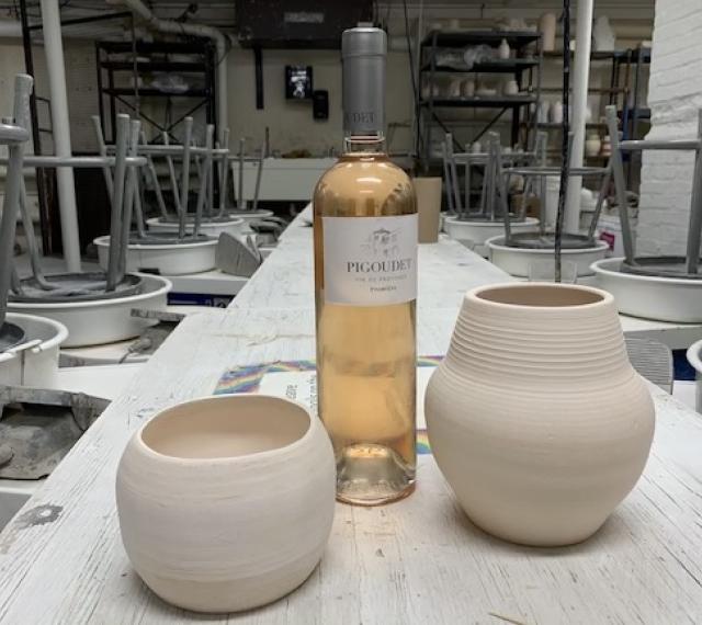 a wine bottle and two handmade pieces of pottery sit in the center table between the rows of pottery wheels in the ceramics studio at MAM.