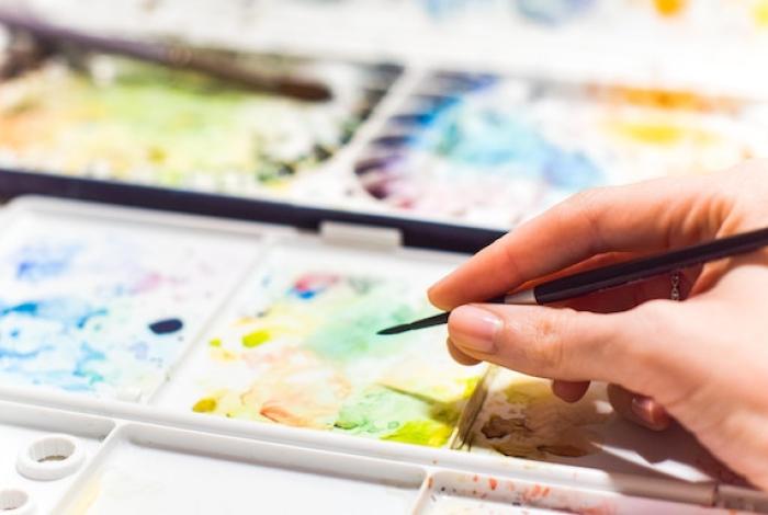 A student is holding a paintbrush, about to dip it into paint on a watercolor palette.